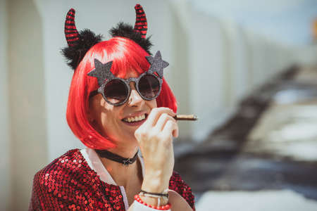 Beautiful girl devil sitting on white background and smoke cigar. In anticipation of Halloween. Closeup Portrait. Wears black sunglassesの写真素材