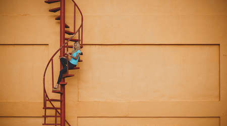 Beautiful leggy blonde is sitting on a red spiral staircase and looking into the distance. On the background of orange walls and sky. Modern street fashion.の写真素材