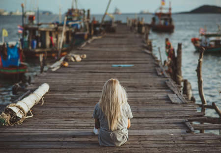 Hipster girl sitting on wooden pier in the rays of the setting sun. A vacation in Thailand. Back to the camera. Vacation in Asia.の写真素材