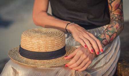 Closeup of a straw hat rests on his knees. Female hand with tattoos correct bracelets. The modern womanの写真素材