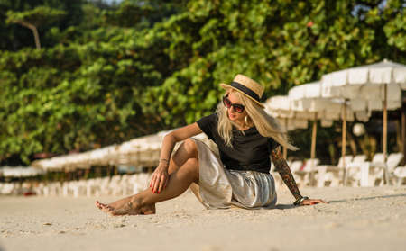 Portrait of a happy young girl. Morning sitting on sandy beach, wearing sunglasses. White umbrellas background. Tattoo modelの写真素材