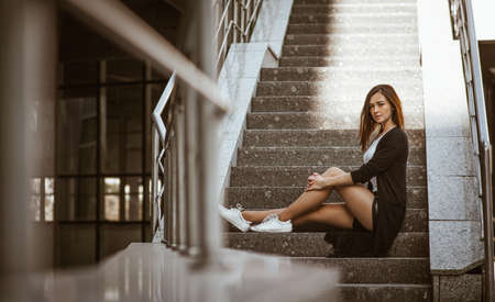 A beautiful girl with red hair wears a black cardigan and short black shorts. Posing on camera sitting on gray urban steps.の写真素材