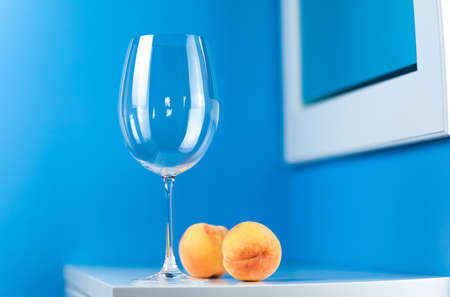 An empty wine glass on a blue background stands on a white table with two peachesの写真素材