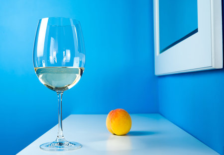 a glass of white wine on a blue background is standing on a white table with a peachの写真素材