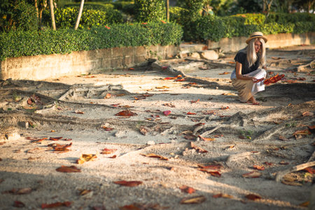 A blonde in a straw hat and long skirt gathers large leaves in a tropical park. Back view. Thailand...