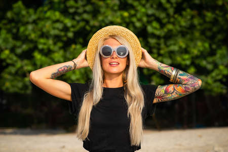 Beautiful blonde is smiling and looking at the morning sea. Holds a hat with his hands with tattoos. Travel to Thailand.の写真素材