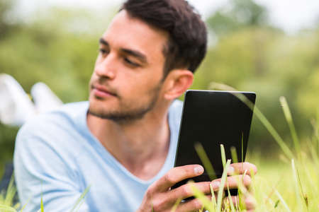 Close-up portrait of young business man with device lying on the green grass and looking at one sideの写真素材