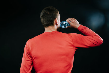 Rear shot portrait of handsome muscular Caucasian jogger wearing red sportswear drinking water,...