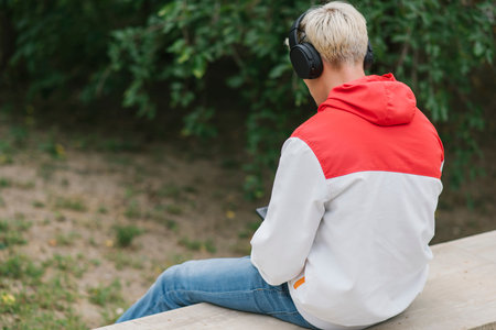 Rear view of young Caucasian blond guy sitting on wall and listening to music outdoors in park....