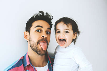 Happy father and beautiful little daughter sticking tongue out, isolated over a white background. Funny faces. Fatherhood. Dad play activities with his girl together shows tongues. Lifestyle family.の写真素材