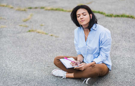 Casual stylish Caucasian student girl sitting at sidewalk, enjoying online communication using wireless internet connection on her smart phone. Young woman with earphone listen and preparing for exam.の写真素材