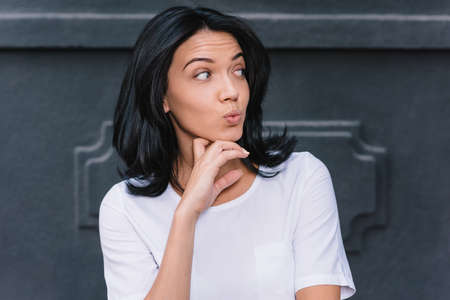 Isolated portrait of joyful young European woman with dark hair touching her chin and looking sideways with surprised expression outdoors with copy space.の写真素材