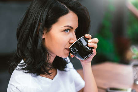 Attractive Caucasian brunette girl dressed in white t-shirt drinking coffee, looking away with serious pensive expression, making plans for day. People and lifestyle concept.の写真素材