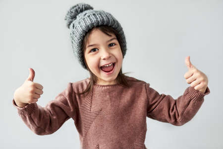 Horizontal closeup portrait of happy smiling little girl in the winter warm gray hat, wearing sweater and showing thumbs up, posing on a white studio background.の写真素材