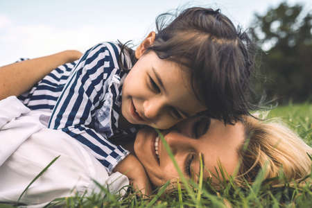 Closeup portrait of young Caucasian woman lying on the green grass and embracing her little daughter in the park. Loving mother and daughter in the park. Happy motherhood time. People and emotionの写真素材