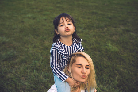 Image of beautiful woman playing and blowing kiss with her cute little girl on green grass outdoor....