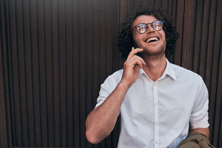 Horizontal image of happy handsome businessman posing against building's wall while standing outdoors and using smartphone for a call.Man with curly hair wearing spectacles talking at his mobile phoneの写真素材