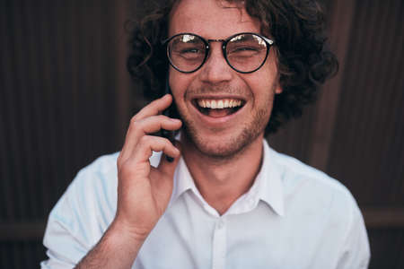 Closeup portrait of happy handsome young businessman making a phone call while walking outdoors wearing white shirt and spectacles. Smiling male with curly hair talking over cell phone on the street.の写真素材