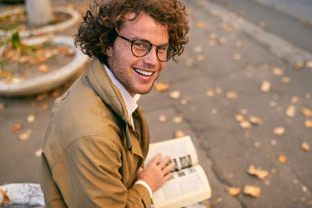 Rear view of happy handsome young man reading book outdoors. College male student carrying books in campus in autumn street. Smiling smart guy wears spectacles and curly hair reading books in streetの写真素材