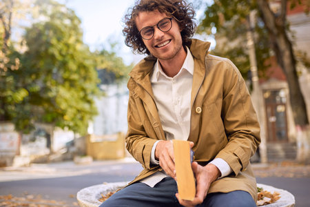 Happy handsome young man reading and posing with book outdoors. College male student carrying books in campus in autumn street. Smiling smart guy wears spectacles and curly hair reading books outsideの写真素材