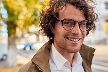 Closeup horizontal portrait of young happy business man with glasses smiling and posing outdoors. Male student in autumn street. Smart guy in casual wears spectacles with curly hair walking on streetの写真素材