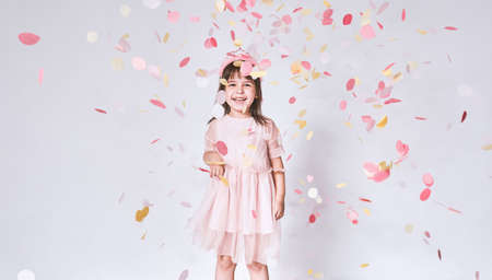 Happy cute little girl wearing pink dress in tulle with princess crown on head isolated on white background playing with confetti. Joyful pretty little girl celebrating her birthday party, having funの写真素材