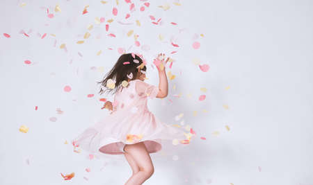 Cheerful cute little girl wearing pink dress in tulle with princess crown dancing on confetti surprise isolated on white studio wall. Playful toddler girl celebrating her birthday party having funの写真素材