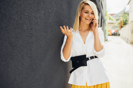 Attractive business woman standing next to gray building's wall while talking on smart phone. Beautiful student female dressed in white shirt, yellow skirt have a call on mobile phone with friend.の写真素材