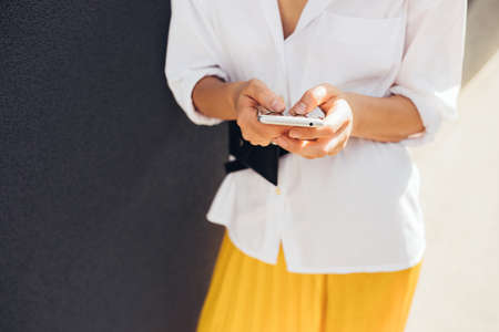 Cropped image of Caucasian young female reading or texting text message on mobile phone on gray wall outdoor in the city. Young woman using free wireless via smart phone on the street. Copy spaceの写真素材