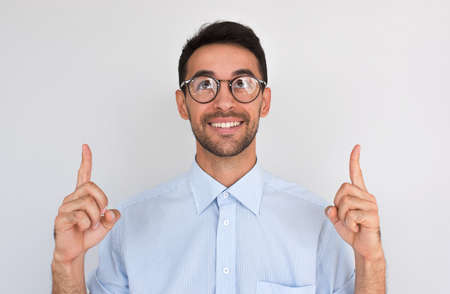Horizontal closeup shot of happy satisfied Caucasian male points upwards with both index fingers, notices something amazing above head, looikng up, wearing round spectacles, against white studio wallの写真素材