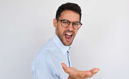 Portrait of angry young handsome male exclaims, feels furious while talking with someone, posing on white studio background. Caucasian man screams nervously, gesture with hand. Negative human emotion.の写真素材