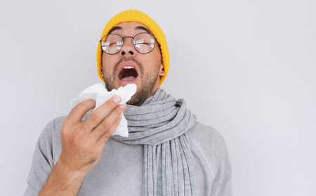 Portrait of sick handsome man wearing grey sweater, yellow hat and spectacles, blowing nose and...