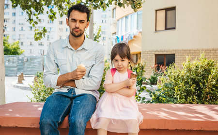 Unhappy cute little girl sitting with dad on the city street and eating ice-cream outdoor. Upset...