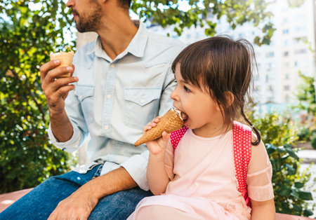 Closeup portrait of happy cute little girl sitting with dad on the city street and eating ice-cream outdoor. Fun girl kid and father have fun outside. Good relationship between dad and daughterの写真素材