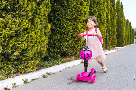 Horizontal shot of happy pretty little girl wearing pink dress and backapack on the scooter against the green trees on the street. Beautiful girl kid preschooler driving scooter in the morning.の写真素材