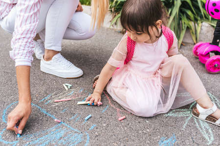 Cropped horizontal image of happy cute little girl and mother drawing with chalks on the sidewalk. Caucasian female play together with kid preschooler with backpack outdoor. Mom and child activityの写真素材