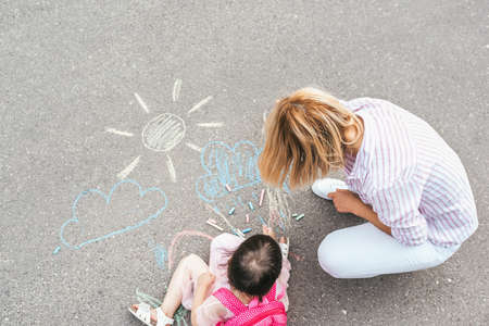 Top view of happy little girl and mother drawing with chalks on sidewalk. Caucasian female play together with kid preschooler with backpack outdoor. Mother and child activity. Good relationshipの写真素材