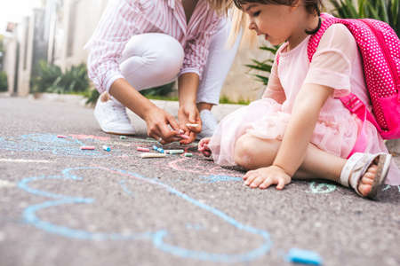 Cropped image of cute little girl and mother drawing with colorful chalks on the sidewalk. Caucasian blonde female play together with her kid preschooler with backpack outdoor. Mom and child activityの写真素材