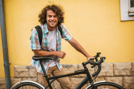 Happy young handsome male with curly hair with backpack, looking to the camera posing with bike on the city street. Man student bicycling to the college. People, sport, lifestyle, healthy life conceptの写真素材