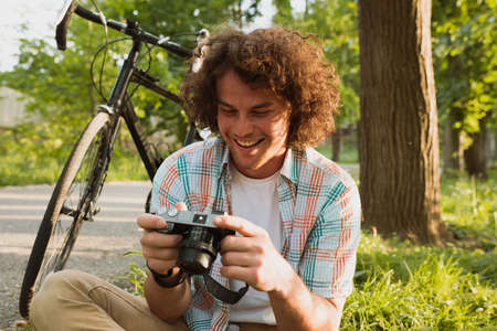 Image of young male smiling with curly hair looking photo on his digital camera. Handsome man wears casual shirt relaxing and sitting near to the bike on mountain. People, travel and lifestyle conceptの写真素材