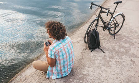 Rear view of young male with curly hair taking photos of nature on his digital camera. Handsome man wears casual shirt relaxing and sitting near to the bike next to the lake. People, travel, lifestyleの写真素材