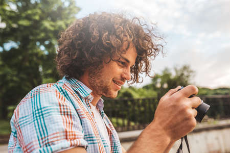 Portrait of Caucasian man with curly hair checking photos in his digital camera. Young handsome male wearing casual shirt, with digital photo camera standing on urban park.People, travel and lifestyleの写真素材