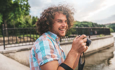 Portrait of Caucasian male with curly hair, taking photos with digital camera on the urban lake. Young man photographing nature and people all around. Happy smiling man exploring with digital cameraの写真素材