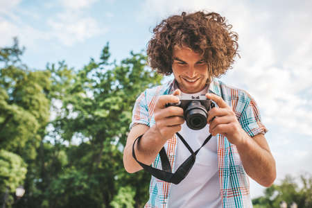 Bottom view shot of Caucasian man with curly hair checking photos in his digital camera. Young handsome male wears casual shirt, with digital photo camera standing on nature background. People, travelの写真素材