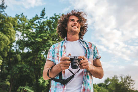 Smiling attractive young tourist man with digital camera taking photos, on a nature and sky background. Handsome man curly hair exploring nature on vacation trip. Travel, people and lifestyle conceptの写真素材