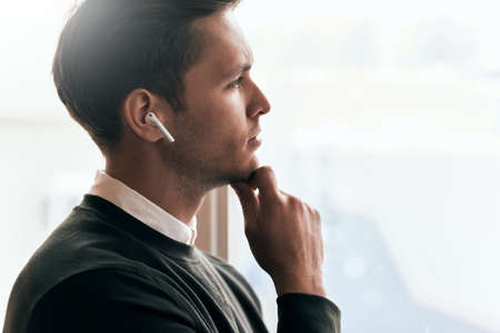 Pensive young handsome man have a call with a colleague and looking through the window in office, talking from wireless earphones. Caucasian businessman using wireless headphones during conversation.の写真素材