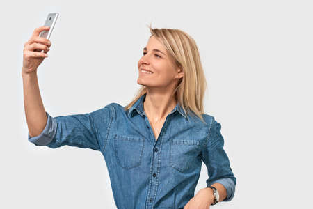 Happy blonde female looks joyfully at camera of smart phone, makes selfie, wearing denim shirt posing against white studio background. Positive student woman taking self portrait on mobile phoneの写真素材