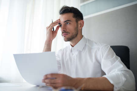 Thinking, working handsome businessman in the office with a paper and a pen in his hand. Business theme. Look ahead with with a hand to his temple, head.の写真素材