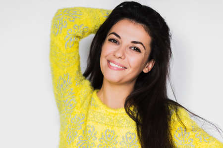 Portrait of brunette cheerful woman with healthy toothy smile, look in front with hands on hair. Young happy pretty woman wearing yellow sweater on light grey background. Positive and happiness mood.の写真素材