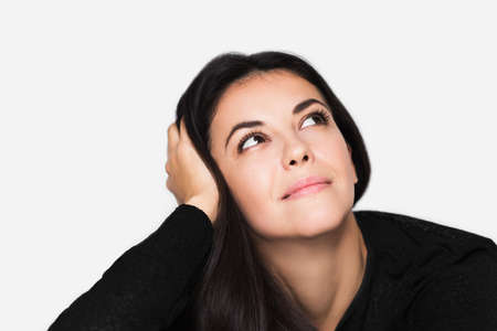 Close up portrait of brunette cute dreamy woman with hand on hair, look up. Young happy pretty woman wearing black sweater on light grey background. Pensive and dreaming mood.の写真素材
